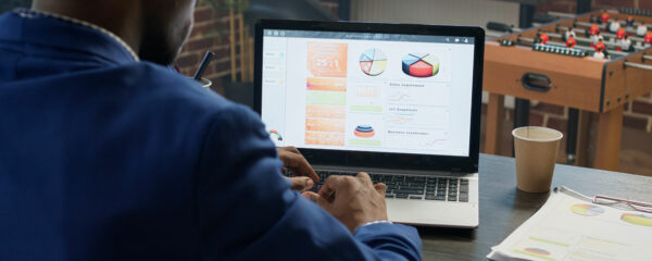 Un professionnel analysant des données et des graphiques commerciaux sur un ordinateur portable dans un bureau moderne. Des documents et un café sont posés sur le bureau, avec un baby-foot en arrière-plan.